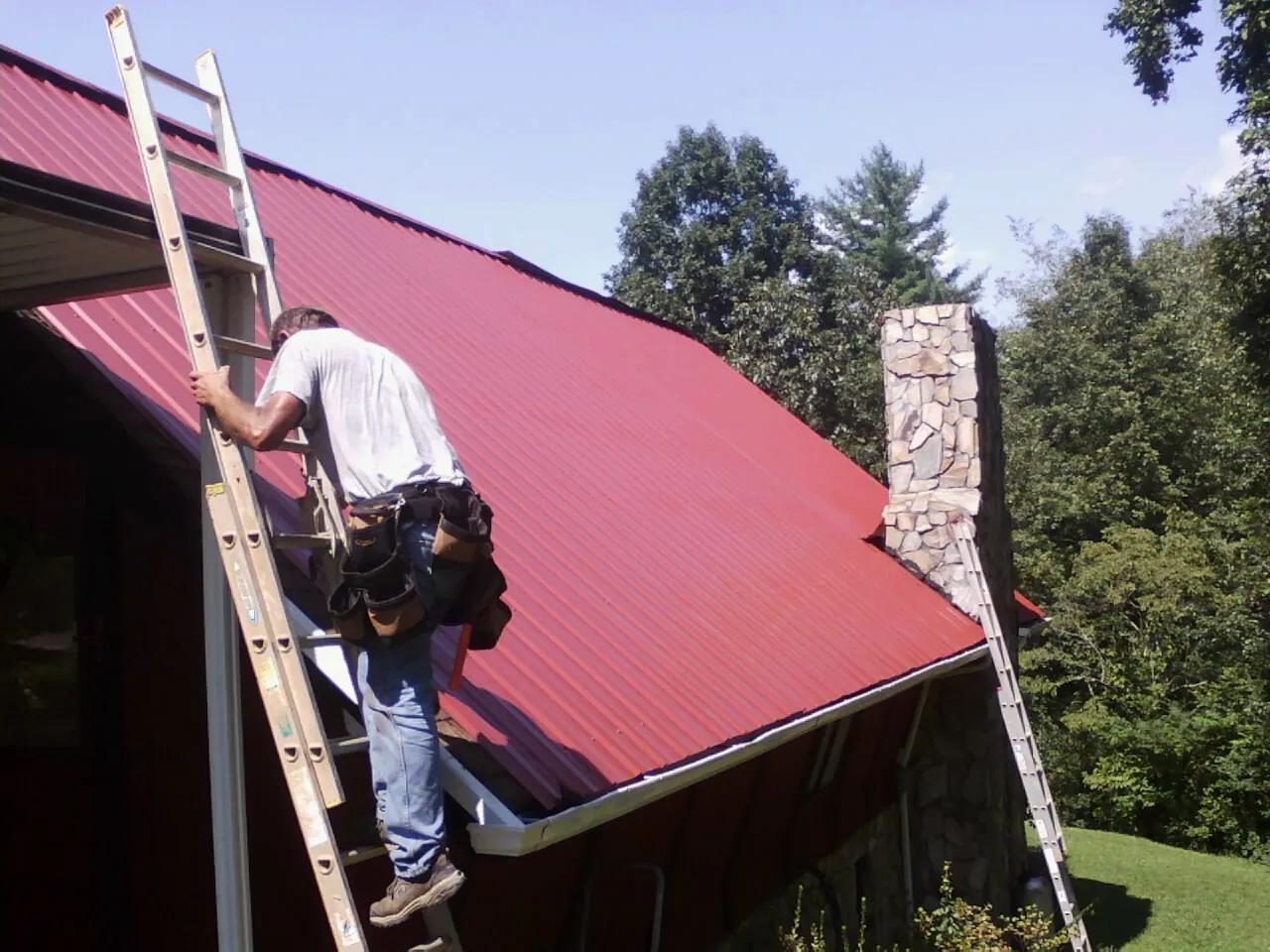 Roofer on a ladder beside a red metal roof and stone chimney — Soffit & Fascia Repair by Roofing Brothers in Hurdle Mills